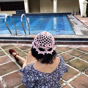 Puede incluir: Un sombrero de ganchillo rosa claro con un dise&ntilde;o calado y peque&ntilde;as cuentas naranjas. El sombrero lo lleva una persona cerca de una piscina. La persona lleva una blusa azul con estampado floral y zapatos rojos.