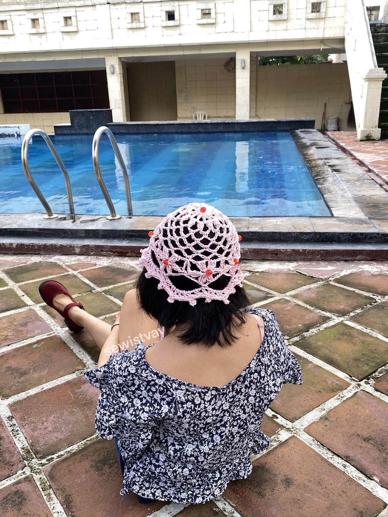 Puede incluir: Un sombrero de ganchillo rosa claro con un dise&ntilde;o calado y peque&ntilde;as cuentas naranjas. El sombrero lo lleva una persona cerca de una piscina. La persona lleva una blusa azul con estampado floral y zapatos rojos.