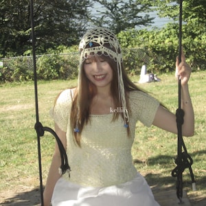Puede incluir: Top de crochet amarillo claro y tocado a juego con detalles azules. El top tiene mangas cortas y un escote cuadrado. El tocado tiene hebras largas y colgantes. El texto "kellii" es visible en la parte superior derecha.