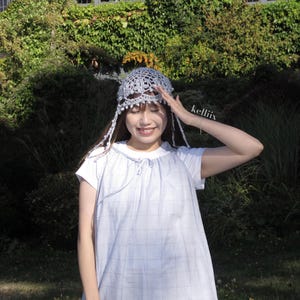 Puede incluir: Vestido azul claro de manga corta con estampado de cuadros. El vestido tiene cuello redondo y detalle de lazo. Se lleva un tocado plateado de ganchillo con hebras colgantes en la cabeza. La imagen est&aacute; tomada al aire libre con un fondo verde.