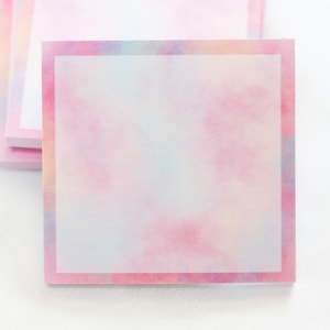 May include: A stack of square sticky notes with a watercolor design. The notes have a soft pink and pastel color scheme with a white center and a pink border. The notes are on a white background.