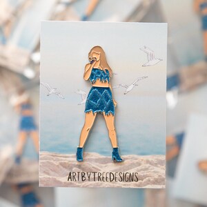 May include: A pin featuring a stylized illustration of a person wearing a blue denim outfit, holding a microphone, with seagulls in the background. The pin has a gold outline and the text "ARTBYTREEDESIGNS" at the bottom.
