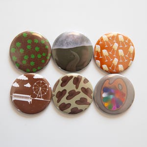 May include: Six round buttons with various designs. One button has green leaves on a brown background. Another features a landscape with a road. Others have champagne glasses, a Ferris wheel, a cow pattern, and a painter's palette.