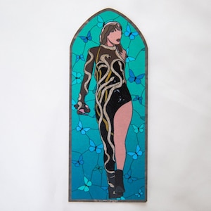 May include: A stained glass artwork featuring a figure in a black bodysuit with gold and white snake-like designs. The background is teal with blue butterflies. The artwork is framed in a dark brown border.