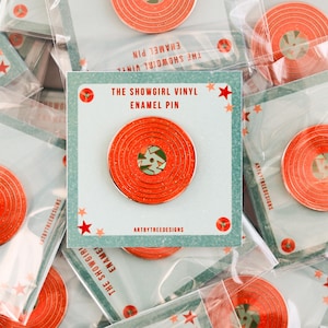 May include: A collection of enamel pins, each depicting a red vinyl record. The pins are packaged on cards with the text "THE SHOWGIRL VINYL ENAMEL PIN." The pins have a glittery finish and a green centre.
