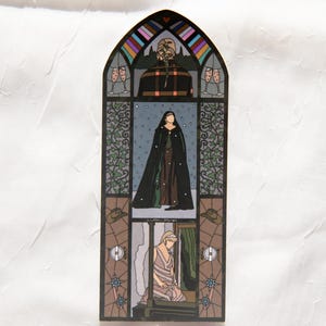 May include: A stained glass art piece with a gothic arch shape. The artwork depicts three scenes: a figure in a plaid coat, a woman in a black cloak, and a person kneeling. The piece features a variety of colors, including black, brown, and blue.