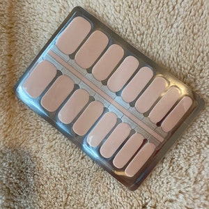May include: A pack of light beige nail polish strips. The strips are arranged in rows within a clear plastic container. The background is a textured, light brown surface.