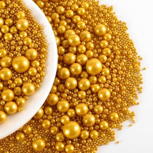 Könnte beinhalten: Nahaufnahme einer weißen Schüssel gefüllt mit goldenen essbaren Perlen, umgeben von einem Haufen ähnlicher Perlen. Die Perlen sind unterschiedlich groß und erzeugen eine strukturierte Darstellung. Die goldene Farbe wirkt luxuriös.