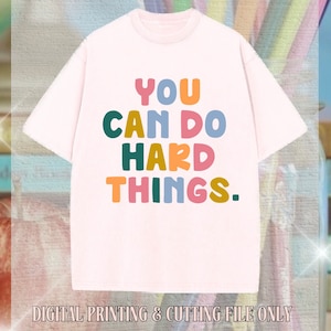 May include: A white t-shirt with the text "YOU CAN DO HARD THINGS." printed in colorful letters. The letters are in different colors, including pink, blue, green, and yellow.