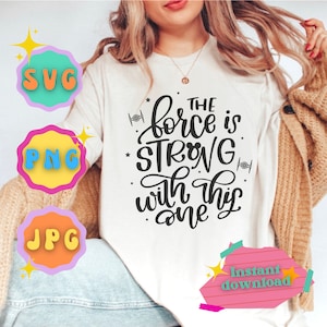 May include: White t-shirt with black text that reads "The force is strong with this one". The text is in a cursive font. There are two small star wars inspired symbols on the shirt. The shirt is being modelled by a person.