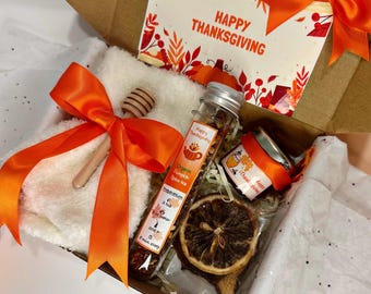 May include: Thanksgiving gift box with an orange bow, containing a white towel, a honey dipper, a tube of organic pumpkin spice tea, a small jar, and a dried orange slice. A card with "Happy Thanksgiving" is visible.