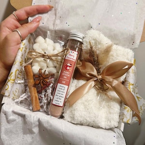 May include: A white gift box filled with a white towel, a glass tube of instant cocoa mix, a bag of marshmallows, a cinnamon stick, and a star anise. The box is decorated with a brown ribbon and a small sprig of dried grass. The cocoa mix tube has a red label with the text "Merry Christmas" and "Instant Cocoa Mix".