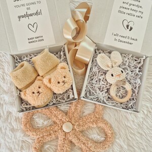 May include: Baby announcement gift set with a teddy bear rattle, bunny rattle, and baby socks. The boxes contain the text "SURPRISE!!! YOU'RE GOING TO BE grandparents!" and "Grandma and grandpa, please keep my little rattle safe."