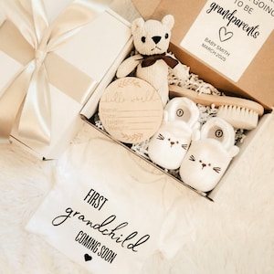 May include: A white gift box with a cream ribbon, containing baby items. Includes a teddy bear, baby shoes, a wooden sign, a baby brush, and a white onesie with the text "FIRST grandchild COMING SOON". A card reads "grandparents".
