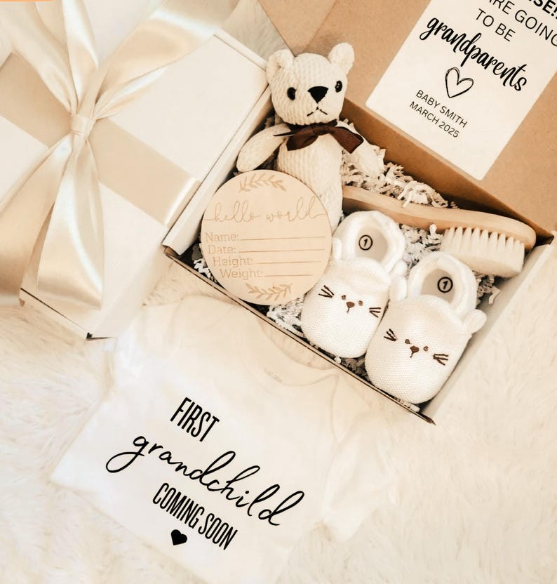 May include: A white gift box with a cream ribbon, containing baby items. Includes a teddy bear, baby shoes, a wooden sign, a baby brush, and a white onesie with the text "FIRST grandchild COMING SOON". A card reads "grandparents".