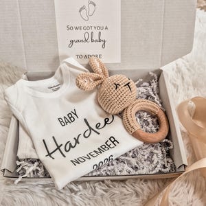 May include: A white Gerber baby onesie with the name "Hardee" and the month "November 2026" printed on it, inside a gift box. A crocheted bunny and wooden ring teether are also in the box. The box also contains a card that reads "We didn't know what to get you, so we got you a grand baby to adore."