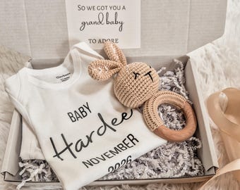 May include: A white baby bodysuit featuring the text Baby Hardee NOVEMBER 2026 and a beige crocheted bunny rattle with a wooden ring. Inside the box, a card reads WE DIDN'T KNOW WHAT TO GET YOU SO WE GOT YOU A grand baby TO ADORE with footprint icons.