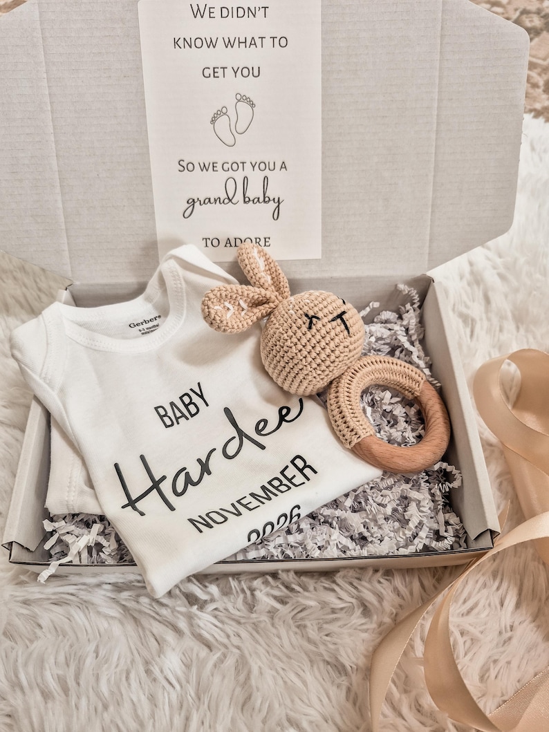 May include: A white Gerber baby onesie with the name "Hardee" and the month "November 2026" printed on it, inside a gift box. A crocheted bunny and wooden ring teether are also in the box. The box also contains a card that reads "We didn't know what to get you, so we got you a grand baby to adore."