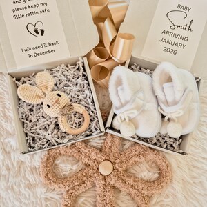 May include: A baby gift set featuring a crochet bunny rattle, white baby booties, and a plush flower-shaped mat. The set includes two gift boxes with text that reads "Grandma and grandpa, please keep my little rattle safe" and "Baby Smith arriving January 2026."