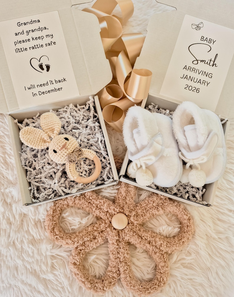 May include: A baby gift set featuring a crochet bunny rattle, white baby booties, and a plush flower-shaped mat. The set includes two gift boxes with text that reads "Grandma and grandpa, please keep my little rattle safe" and "Baby Smith arriving January 2026."