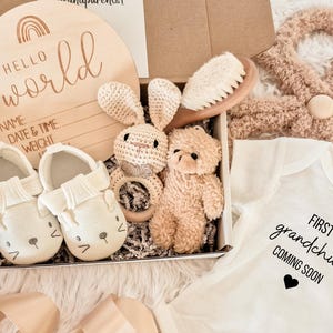 May include: A baby gift set in a box, including a wooden sign with "Hello World" and lines for name, date, and weight. The set also contains baby shoes, a crochet bunny rattle, a teddy bear, a baby brush, and a onesie that says "First grandchild coming soon."