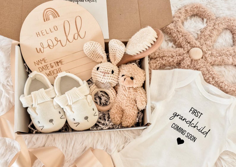 May include: A baby gift set in a box, including a wooden sign with "Hello World" and lines for name, date, and weight. The set also contains baby shoes, a crochet bunny rattle, a teddy bear, a baby brush, and a onesie that says "First grandchild coming soon."