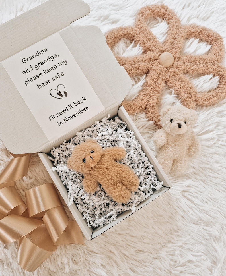 May include: A small, tan teddy bear rests in an open cardboard box filled with white paper shreds. The box contains a note that reads, "Grandma and grandpa, please keep my bear safe. I'll need it back in November." A larger, tan, flower-shaped item and a smaller teddy bear are also visible.