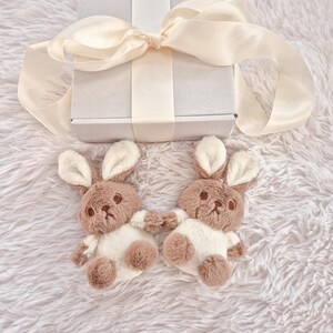 May include: Two plush bunny toys with brown and white fur, positioned on a fluffy white surface. The bunnies are holding hands. A white gift box with a cream-colored ribbon is in the background.