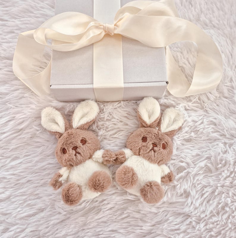 May include: Two plush bunny toys with brown and white fur, positioned on a fluffy white surface. The bunnies are holding hands. A white gift box with a cream-colored ribbon is in the background.