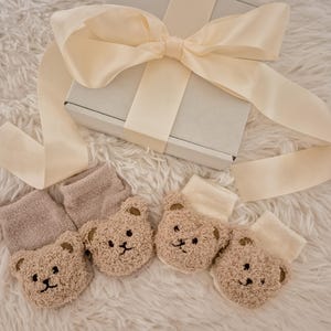 May include: A gift set featuring a light gray box tied with a cream-colored ribbon. Four pairs of teddy bear-faced socks in shades of beige and cream are arranged on a fluffy white surface, creating a cozy and gift-ready presentation.