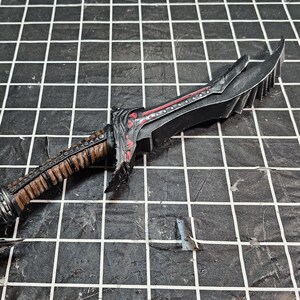 May include: A detailed, black dagger with a serrated blade and a brown and black handle. Red accents are visible on the blade and handle. The dagger is set against a black and white grid background.