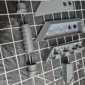 Puede incluir: Vista cenital de varios componentes grises impresos en 3D, posiblemente para un modelo o prototipo. Las piezas incluyen formas cilíndricas y rectangulares, junto con piezas con agujeros y bordes angulados. Dispuestas sobre una superficie de cuadrícula en blanco y negro.