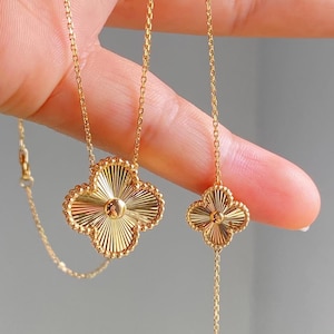 Puede incluir: Juego de collar y pulsera dorados con forma de trébol. La joyería presenta un borde con cuentas y un diseño de rayos de sol en los colgantes de trébol. El collar y la pulsera se sostienen sobre un fondo neutro.