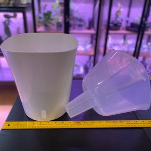 May include: A white, self-watering planter with a clear plastic insert. The planter has a rectangular shape with rounded corners. A yellow ruler is placed next to the planter. The planter is designed for indoor use.