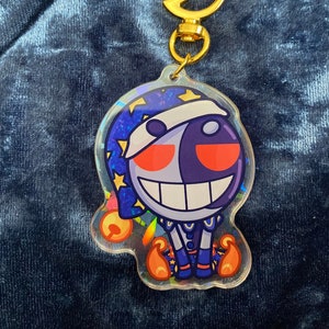 May include: A holographic charm featuring a cartoon character with a white and purple face, red eyes, and a large smile. The character is wearing a blue and white outfit and a blue hat with stars. The charm is attached to a gold crescent moon keychain.