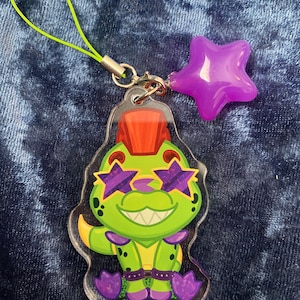May include: A green cartoon dinosaur with a mohawk, wearing purple star-shaped sunglasses, is featured on a clear acrylic keychain. The keychain has a lime green lanyard and a purple star charm.