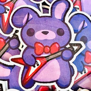 May include: A purple plush toy rabbit with a red bow tie is holding a red electric guitar. The toy is a sticker with a white outline.