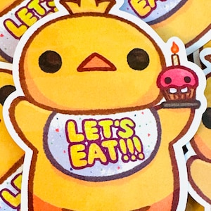 May include: A yellow chick with a crown and a bib that says "Let's Eat!!" is holding a pink cupcake with a lit candle. The chick has big black eyes and a small orange beak.