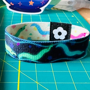 Könnte beinhalten: Ein schwarz-weißes Armband mit einem bunten Design mit einer Blume und wellenförmigen Linien. Das Armband hat eine blaue, grüne und rosa Farbgebung.