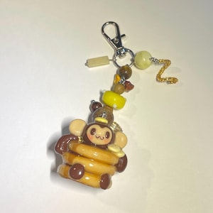 May include: A keychain featuring a brown monkey character sitting atop a stack of golden-brown pancakes. The charm is attached to a silver-toned lobster clasp with a decorative arrangement of yellow, brown, and clear beads, plus a gold-toned chain.