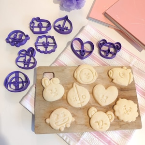 Puede incluir: Cortadores de galletas de plástico morado en varias formas, incluyendo corazones, estrellas y animales, están dispuestos en una superficie blanca. Junto a los cortadores hay varias galletas cortadas en las mismas formas, dispuestas en una superficie con estampado blanco y rosa.