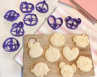 May include: Purple plastic cookie cutters in various shapes, including hearts, stars, and animals, are arranged on a white surface.  Next to the cutters are several cookies cut out in the same shapes, arranged on a white and pink patterned surface.