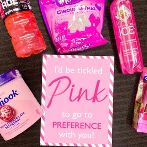 May include: A pink and white striped card with the text "I'd be tickled pink to go to PREFERENCE with you!"  Surrounding the card are various pink-themed snacks and drinks, including a pink sports drink, a pink ice cream container, a bag of pink cookies, a pink sparkling water bottle, and a box of pink chocolate squares.