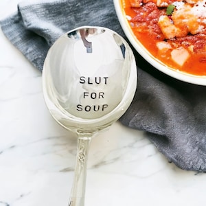 May include: A silver soup spoon with the words "Slut for Soup" engraved on the bowl. The spoon is sitting on a gray napkin next to a bowl of soup.