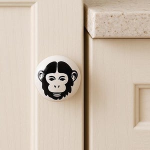 May include: A white cabinet knob featuring a black monkey face design. The round knob has a glossy finish. The monkey face is a simple, stylized illustration. The knob is attached to a light-coloured cabinet door.