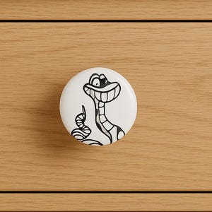 May include: A white ceramic drawer knob featuring a black cartoon snake design. The knob is attached to a light brown wooden drawer. The snake has a smiling face and is pointing upwards. This decorative knob is ideal for adding a touch of fun to furniture.