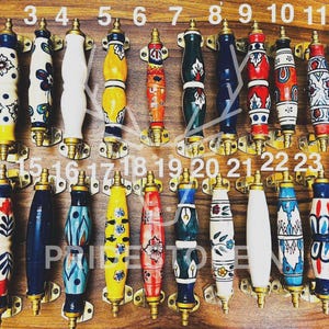 May include: A collection of twenty-four colorful ceramic cabinet pulls with brass hardware. Each pull features unique hand-painted designs in various colors, including blue, red, yellow, and white. The pulls are arranged on a wooden surface.