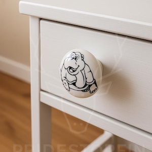 May include: A white ceramic drawer knob with a black outline cartoon hippo design. The knob is fixed to a white painted wooden drawer, part of a white painted cabinet.