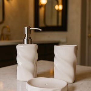 May include: A white ceramic bathroom set featuring a soap dispenser with a silver pump, a tumbler, and a soap dish. Each piece has a textured, twisted design. The set is displayed on a marble surface.