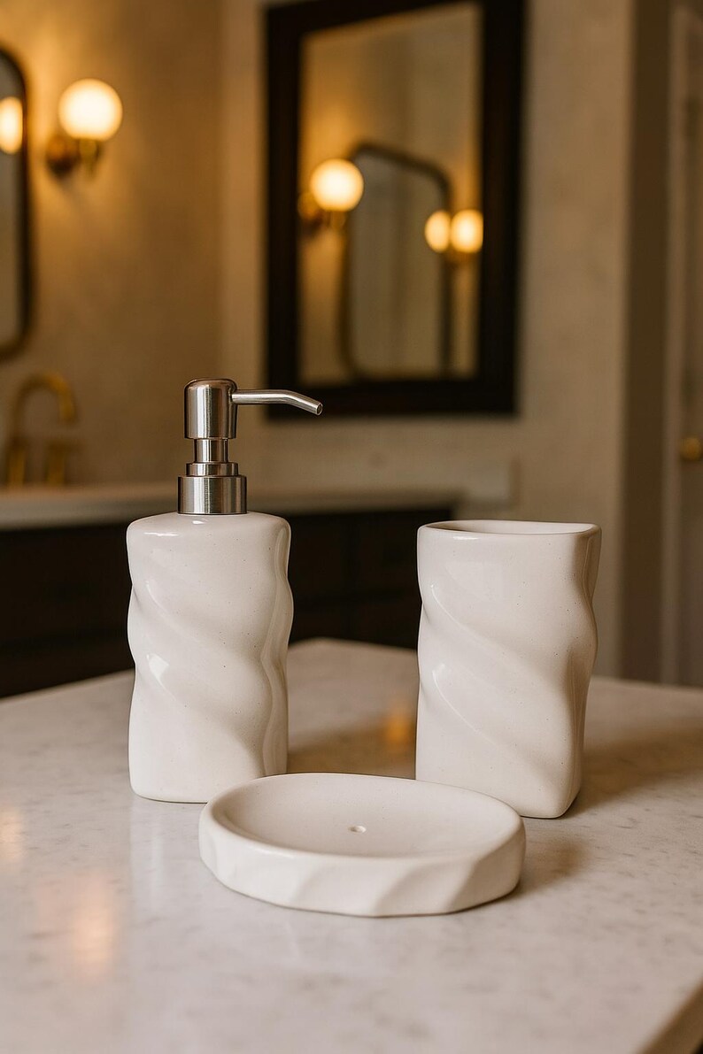May include: A white ceramic bathroom set featuring a soap dispenser with a silver pump, a tumbler, and a soap dish. Each piece has a textured, twisted design. The set is displayed on a marble surface.